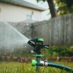 A rotating garden sprinkler irrigating a backyard lawn with a fence and house in the background.